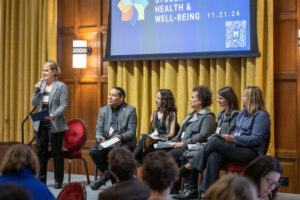 A moderator talks into a microphone while a man and four women sit and look on.