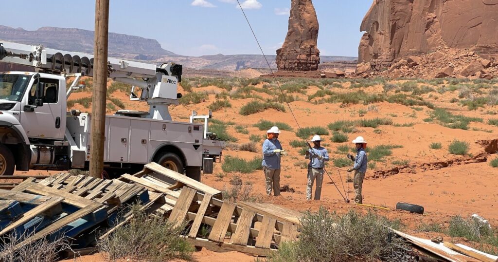 Five electric crew workers and a woman whose home they connected to the electric grid stand in a group photo in the desert. In the background is her trailer home and red rock mesas.