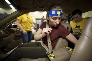 A woman, wearing a sensor strip across her forehead straps into the passenger seat of a test vehicle while a researcher in the back seat works on a laptop computer.