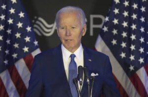 A man in a blue suit and tie speaks at a podium with microphones, standing in front of two American flags and a dark background with partial text visible.