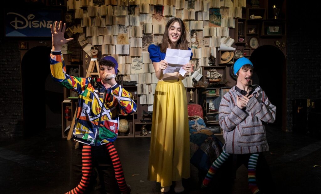 A man with a beard stands outdoors on a sunny day, wearing a brown jacket. Behind him are blooming trees, a grass lawn, a path, and brick buildings.