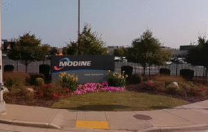 Exterior view of a sign reading Modine World Headquarters, surrounded by landscaped flowers and bushes on a sidewalk near a parking lot.