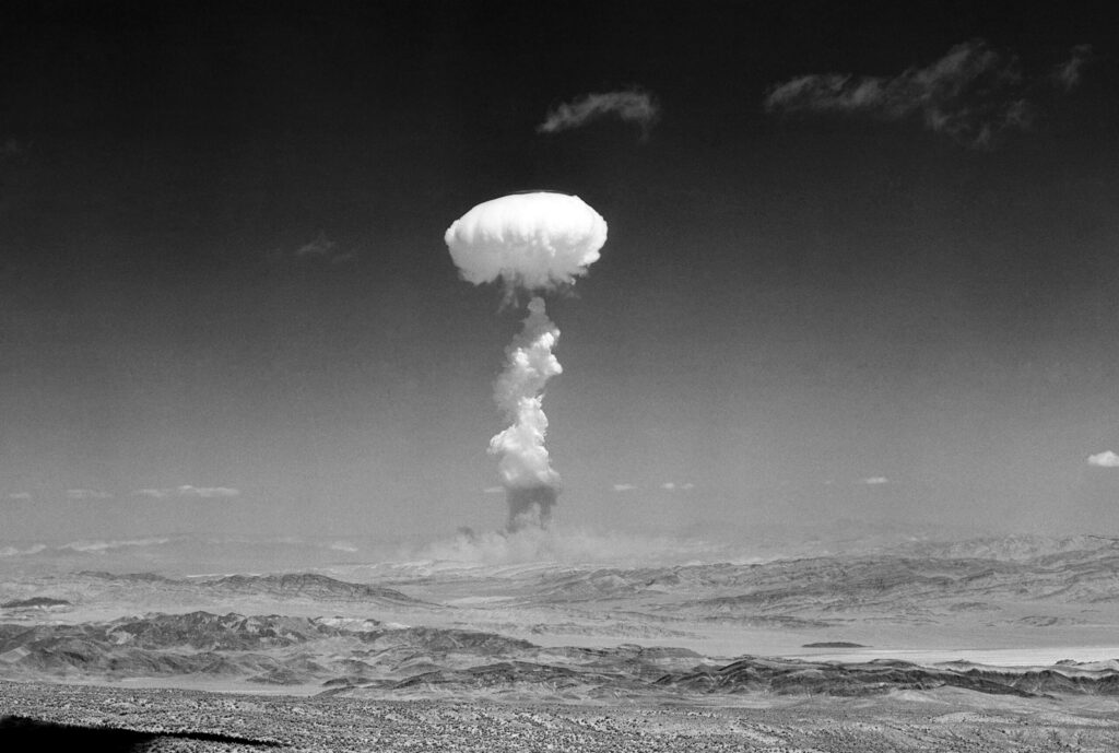 A large mushroom cloud rises into the sky over a desert landscape, indicating the aftermath of an explosion or detonation.
