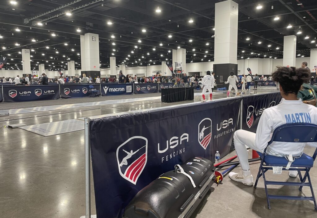 Two fencers in protective gear compete on a fencing strip at an indoor tournament with USA Fencing banners visible.