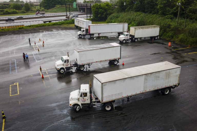 Aerial view of several white semi-trucks with trailers maneuvering in a wet parking lot, with a few people standing nearby and highways visible in the background.