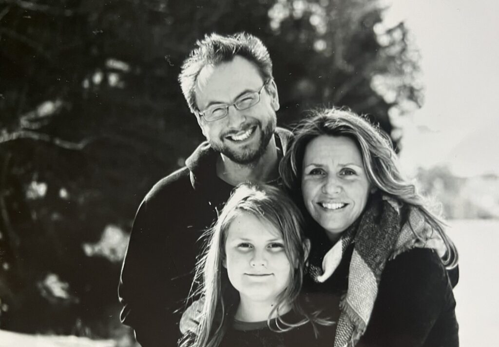 An adult and a child, both wearing dark jackets, smile while posing together in front of tall green corn stalks outdoors.