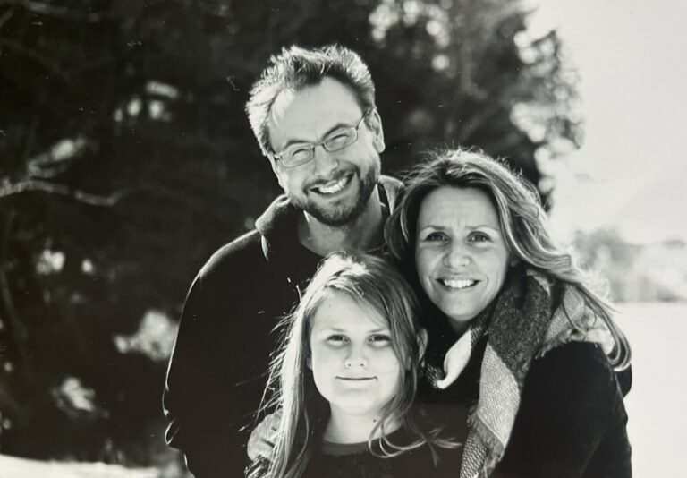 An adult and a child, both wearing dark jackets, smile while posing together in front of tall green corn stalks outdoors.