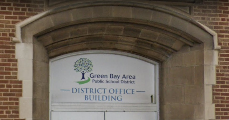 A man in a suit and tie is smiling, wearing a necklace with a rectangular pendant, posing against a dark, blurred background.