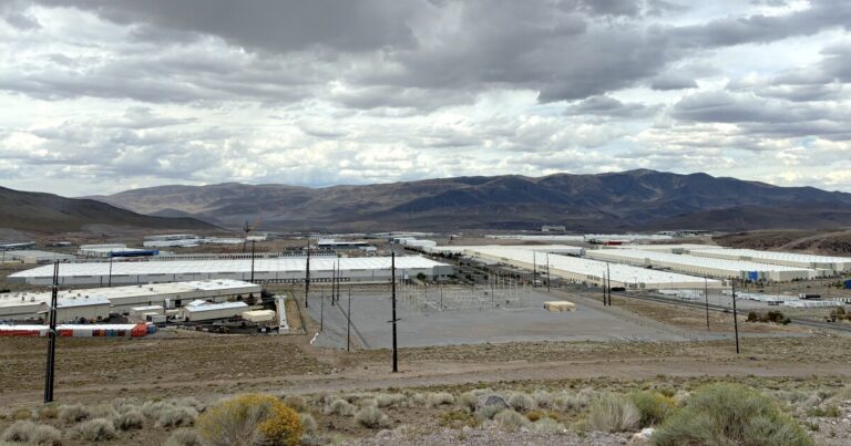 On the right of the image, in the foreground, is a white sign with 'Google' on it. In the background is a brown building that is the company's data center, backdropped by a clear blue sky and mountains.