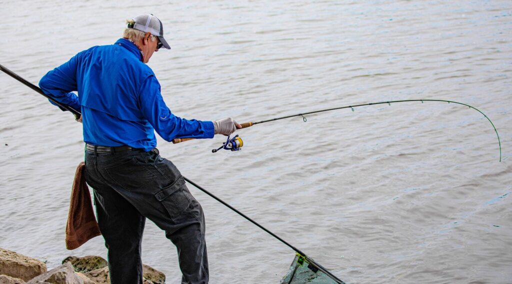 Two men standing by the water, smiling and holding a large fish. One gives a thumbs up. Industrial buildings and a grassy area are visible in the background.