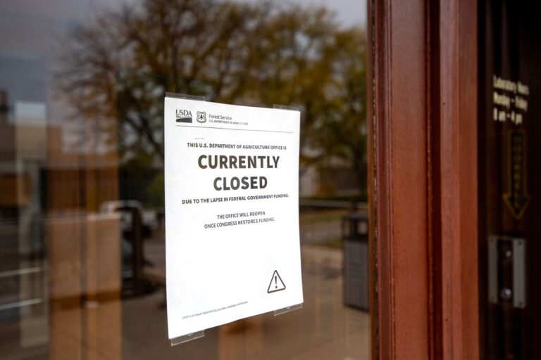 A government building with multiple floors, an American flag on the roof, several cars parked in front, and trees on either side of the entrance.