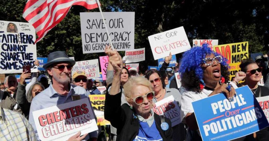 House Speaker Destin Hall leans over his wooden lectern at the front of the House floor to speak to a man. Behind him is the North Carolina state seal in gold. He wears a light pinstriped suit with a navy pocket square.