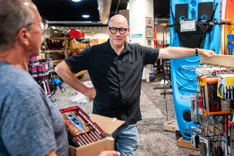 Two men converse in an outdoor gear store; one is holding a box, while the other stands with his hand on his hip near displayed kayaks and camping equipment.