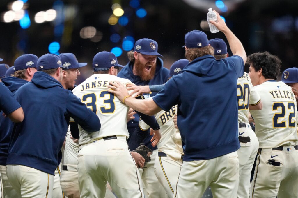 A Milwaukee Brewers baseball player in uniform yells in celebration on the field, clenching his fist, while an umpire stands in the background.