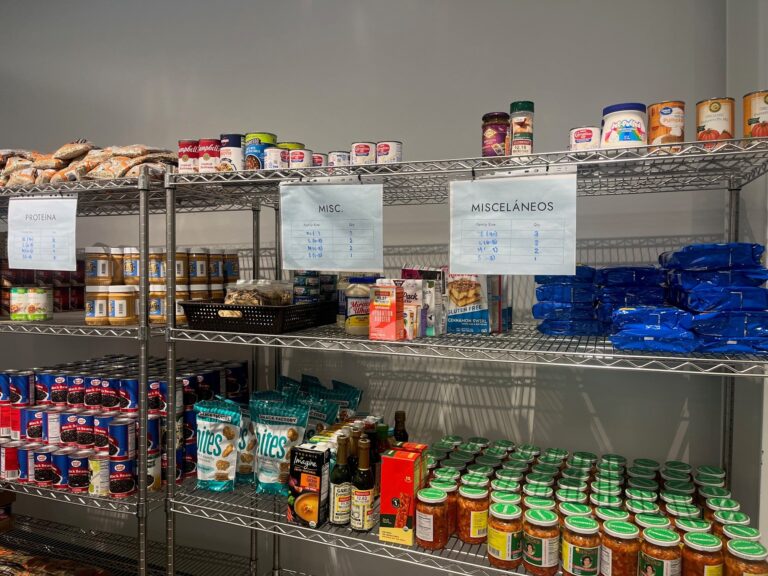 Metal shelves stocked with various non-perishable foods, including pasta, canned goods, snacks, and sauces. Signs above the shelves label the sections and list item limits.