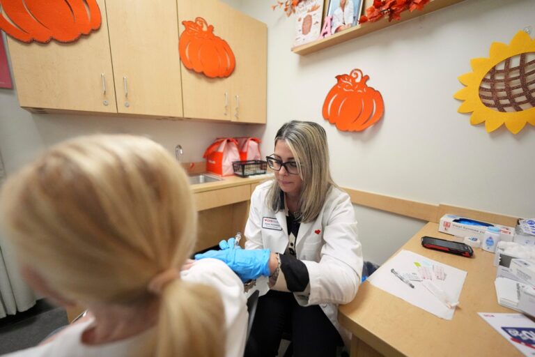 A healthcare worker in a white coat prepares to administer a vaccine to a seated patient in a decorated clinic room.