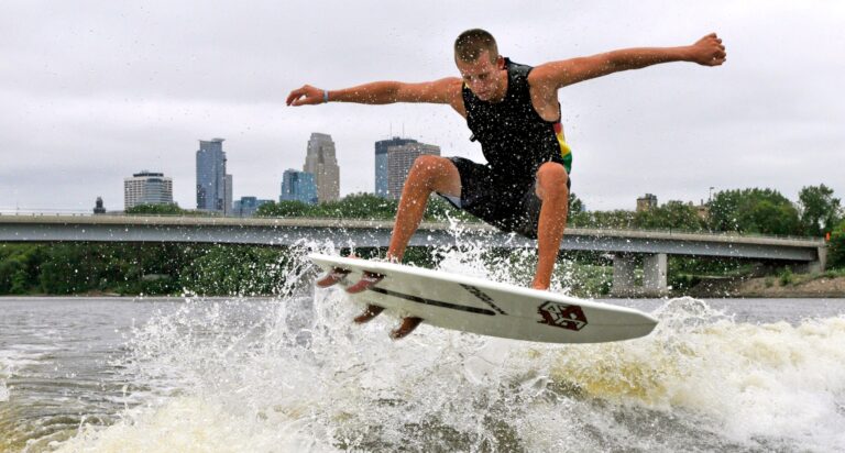 A person in a black tank top is wakesurfing and airborne above the water, with city buildings and a bridge in the background.