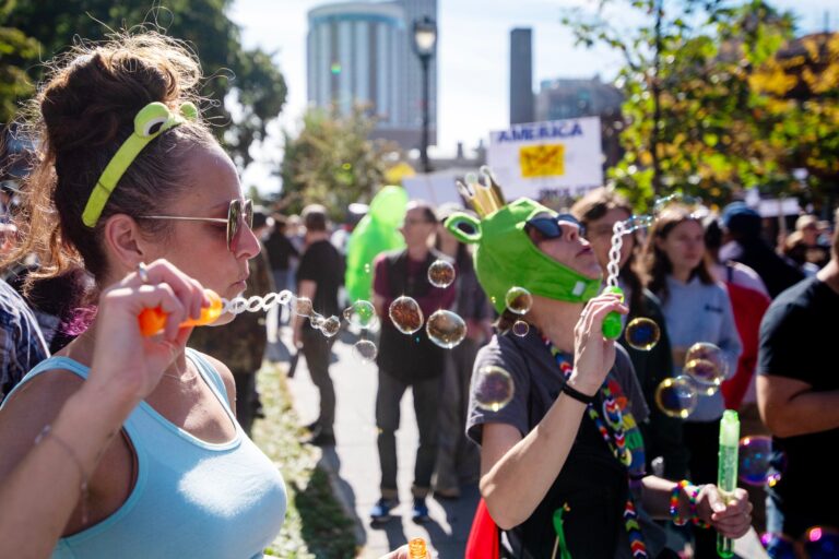 A group of people march down a city street holding protest signs, including one reading NOPE NOT IN WISCONSIN, with buildings visible in the background.
