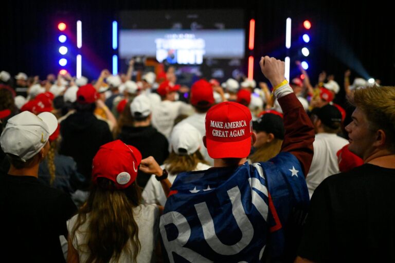 A group of people sit around a conference table with American flags, watching a projected video of a man giving a speech on a screen at the front of the room.