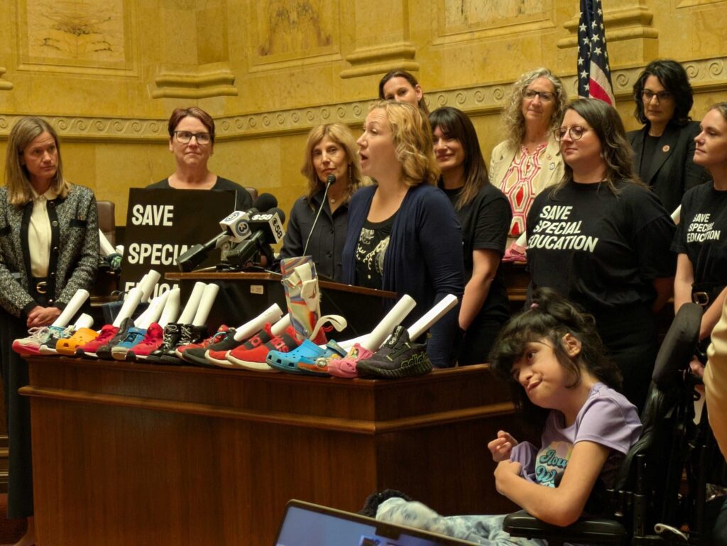 A group of women, some in Save Special Education shirts, stands at a podium with childrens shoes and advocacy signs; a girl in a wheelchair is in the foreground.