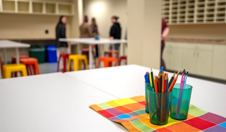 A classroom bulletin board displays various math problems and numbers on colorful cards, with string lights visible along the edge.