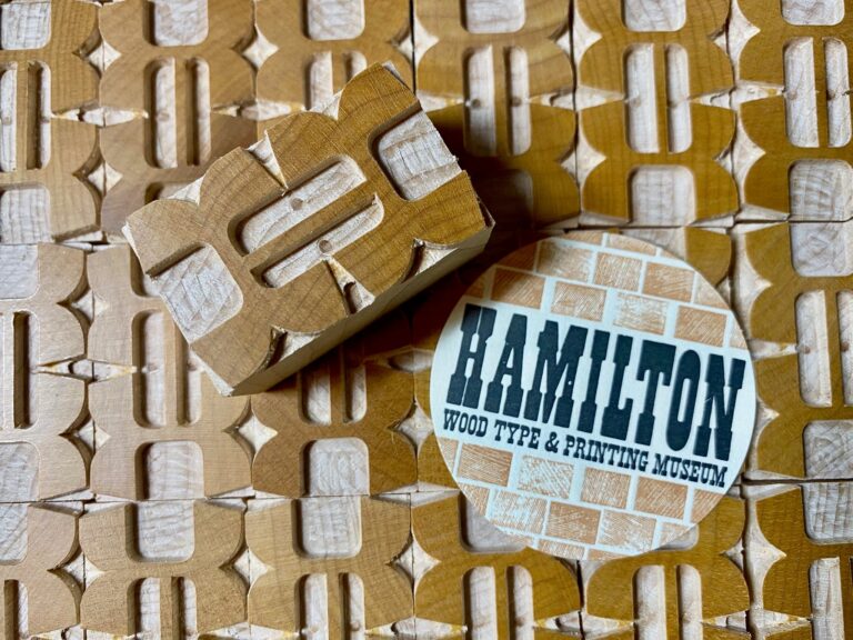 A wood type printing block and a circular sticker reading Hamilton Wood Type & Printing Museum on a patterned wooden surface.