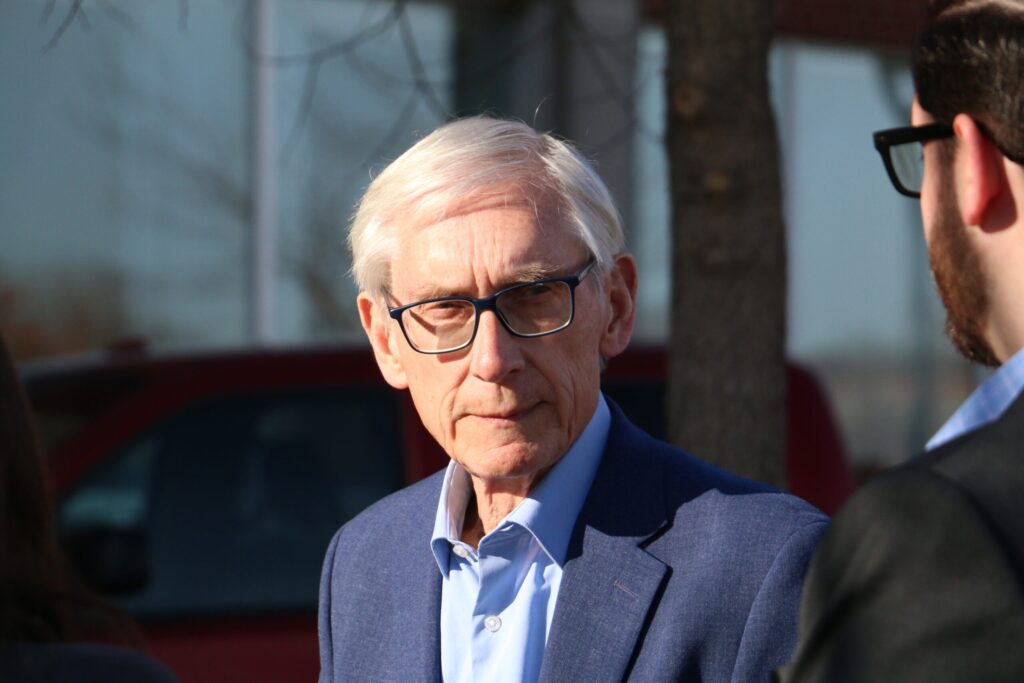 An older man with white hair and glasses wearing a blue suit jacket stands outdoors, facing another person.