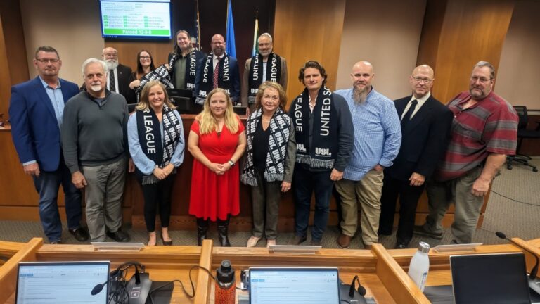 A group of people stands in a government meeting room, some wearing black and white United Soccer League scarves, with desks and monitors in the foreground.