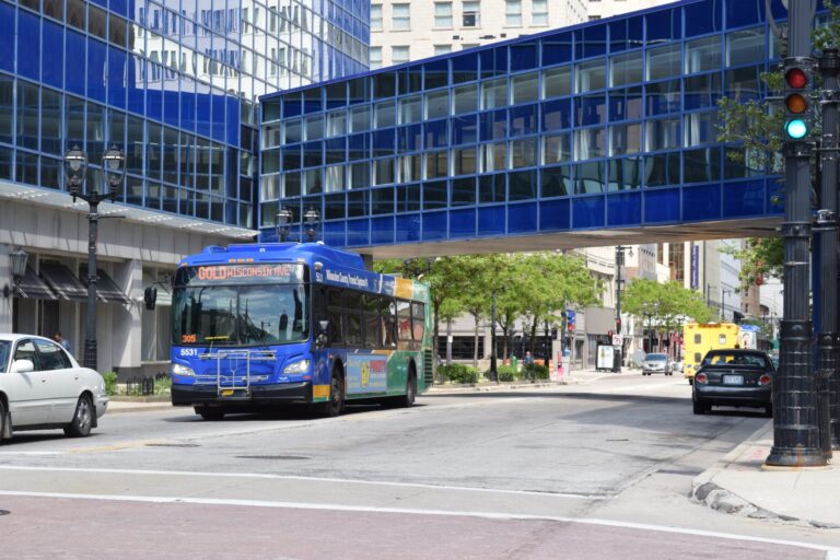 A city bus marked Green MEGA stops at a bus shelter on an urban street, with people boarding and buildings in the background.