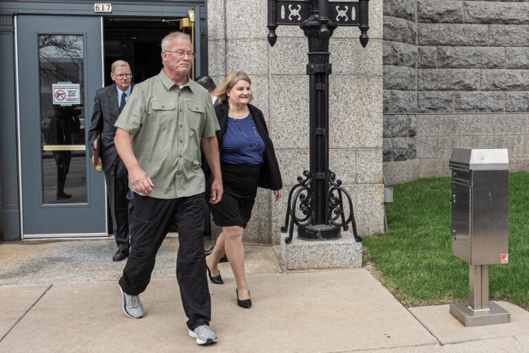 Three adults walk out of a building with stone walls, one man in front in casual clothes, a woman in business attire behind him, and another man in a suit exiting the door.