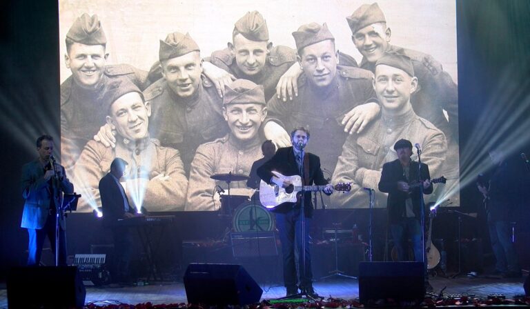 A band performs on stage in front of a large projected black-and-white photo showing a group of smiling soldiers in uniform.