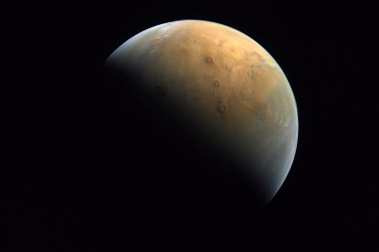 A partially shadowed view of Mars shows surface features and craters, with the planet’s reddish-brown terrain visible against the dark background of space.