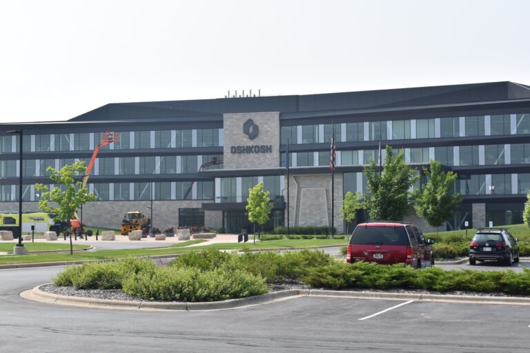 Modern office building with Oshkosh signage on facade, several vehicles parked in front, and construction equipment near entrance, surrounded by landscaping.