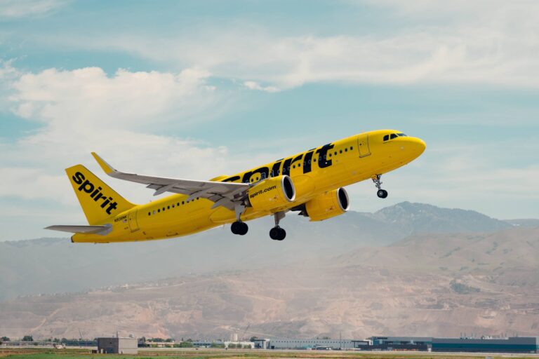 A bright yellow Spirit Airlines airplane takes off from a runway with mountains and clouds in the background.