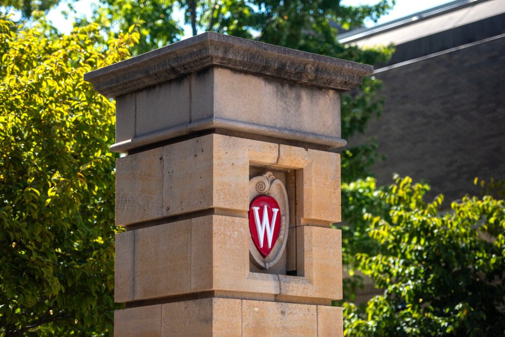 A stone pillar with the University of Wisconsin crest featuring a red W is shown against a background of trees and a building.