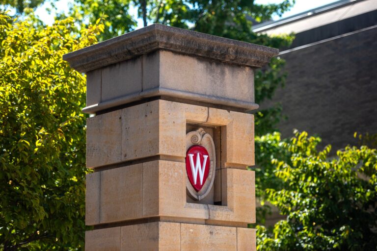 A stone pillar with the University of Wisconsin crest featuring a red W is shown against a background of trees and a building.