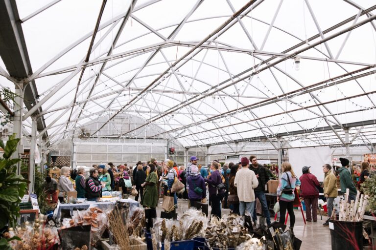 A crowd of people browse stalls with plants and goods inside a spacious greenhouse with a glass ceiling.