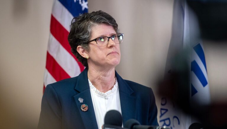 A person stands at a podium in a room, speaking at a press conference, surrounded by cameras, microphones, and information boards.