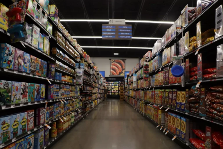 People browse shelves stocked with canned goods and soups in a crowded food pantry. Signs above the shelves label sections such as Soups + Ready to Eat and Bread.
