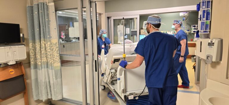 Medical staff in scrubs move a patient on a hospital bed through a glass door in a hospital ward.