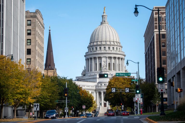 Street view of the Wisconsin State Capitol building with its white dome, flanked by city buildings and autumn trees under a clear blue sky.