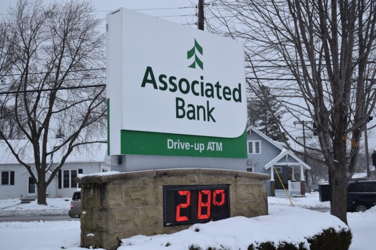 A beige stone bank building with a snow-covered roof and surrounding bushes, with two cars parked in front on a snowy, overcast day.