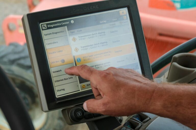 A person’s hand operates the touchscreen diagnostics center of a John Deere machine, displaying system information and troubleshooting options.
