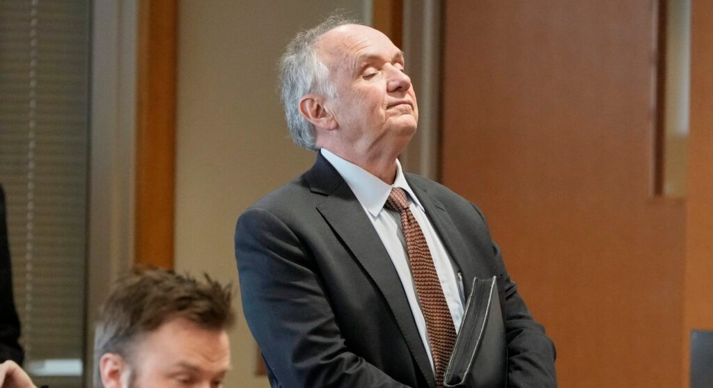 An older man in a suit stands in a courtroom holding a folder, while another man sits nearby looking down.