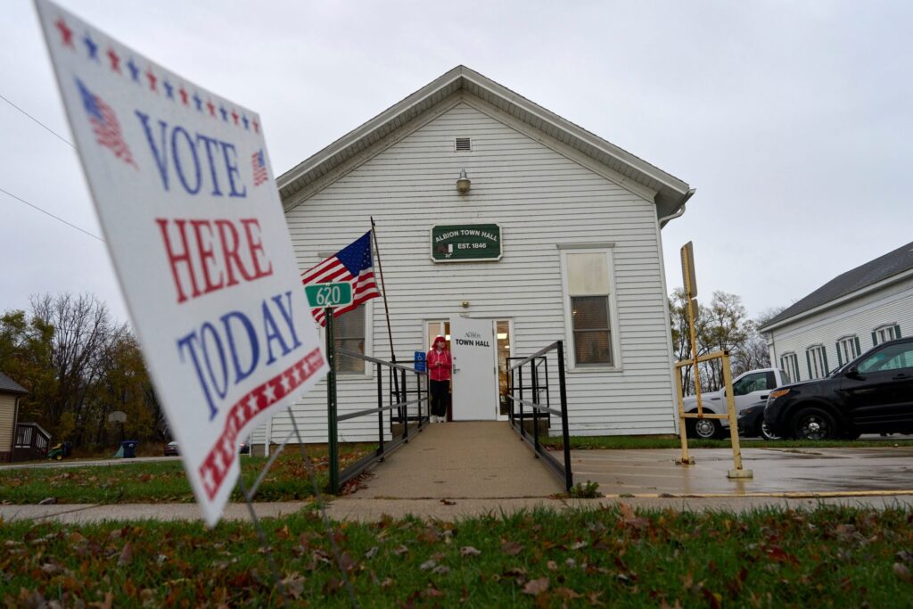 A sign reading Vote Here Today stands outside a white building with a ramp and an American flag; a person is entering the building.