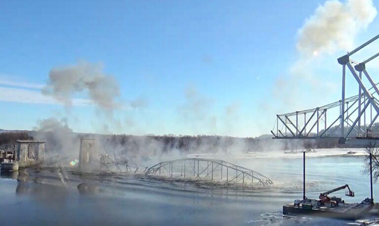 A woman wearing a white knit hat and a Black Hawk Bridge Lansing, Iowa hoodie stands indoors, smiling at the camera.