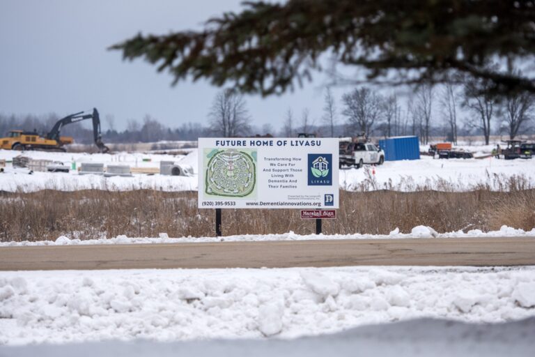 A snow-covered construction site with visible building foundations, a few vehicles, and portable office structures, surrounded by roads and open land.