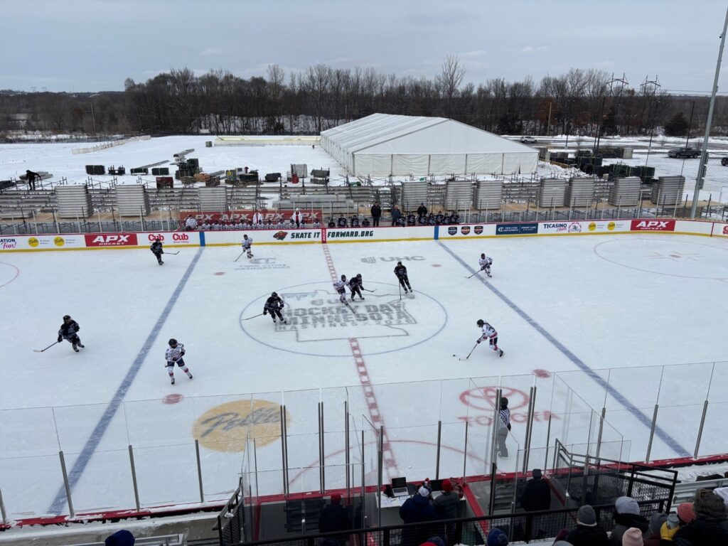 An empty outdoor hockey rink with surrounding bleachers and lit stadium lights at dusk, with a cloudy sky and trees in the background.
