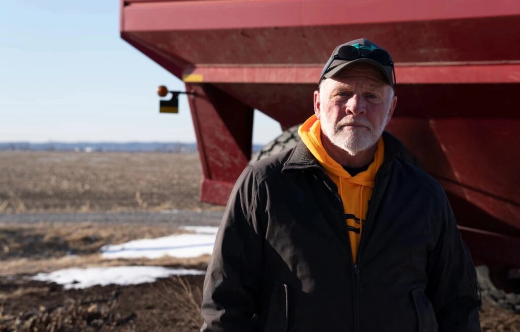 An older man in a black jacket and orange hoodie stands outdoors near a large red farm vehicle on a partly snow-covered field.