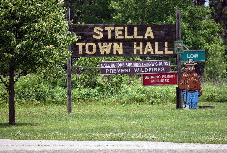 Large Stella Town Hall sign with wildfire prevention notices and a Smokey Bear cutout beside a green Low fire danger sign, set in a grassy area with trees.